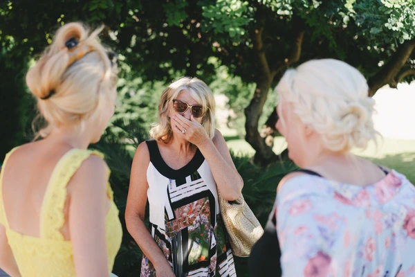 Female guests chatting under green trees on manicured lawn — woman in sunglasses gesturing