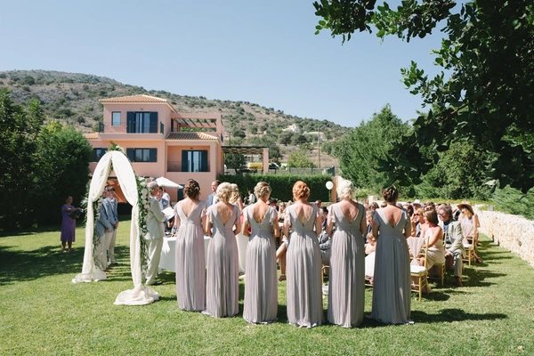 Light grey bridesmaid dresses looking toward the altar — white floral arch, sunny Mediterranean villa
