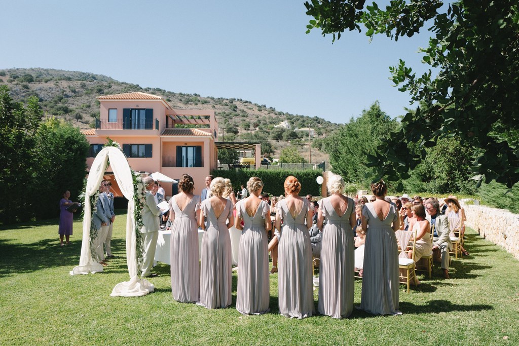 Light grey bridesmaid dresses looking toward the altar — white floral arch, sunny Mediterranean villa