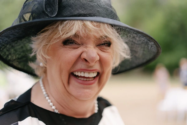 Elderly woman laughing joyfully wearing black hat with pearl necklace at garden wedding celebration.