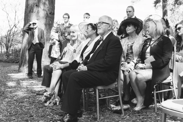 Black and white candid shot of seated wedding guests including parents and family members watching the ceremony intently. Documentary audience moment.