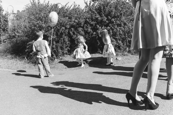 Black and white candid shot of three children casting long shadows on the road as they play with a balloon in the sunlit garden. Documentary childhood moment at a wedding.