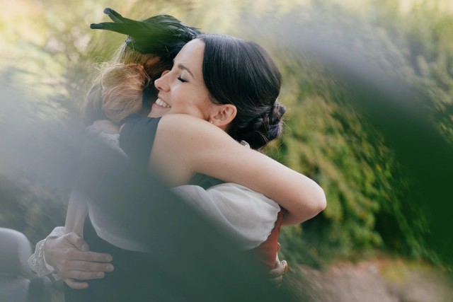 Smiling dark-haired bride shares joyful embrace with guest at outdoors wedding in soft golden light