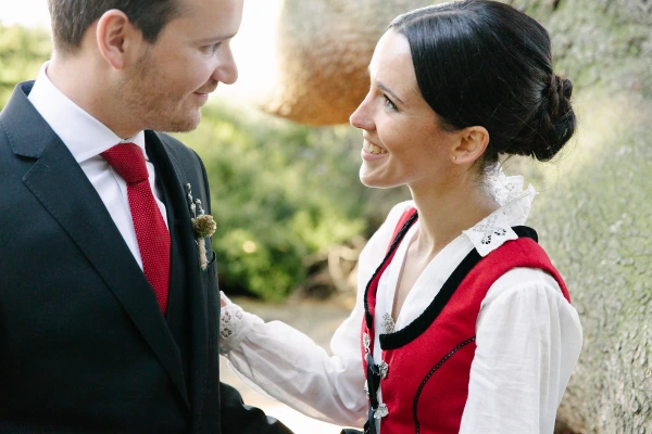 Bride and groom smiling at each other while leaning against a tree in the Royal Botanic Garden Edinburgh, bride in Norwegian bunad and groom in dark suit with red tie. Warm couple portrait.