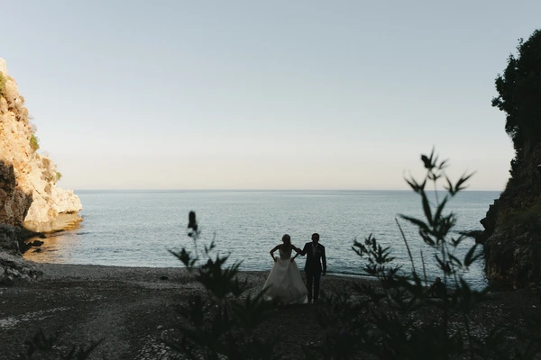 Silhouette of bride and groom on the beach — calm sea, clear sky, soft light, blurred plants