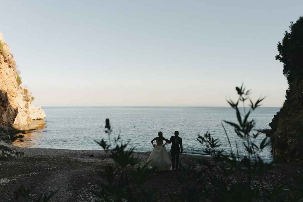 Silhouette of bride and groom on the beach — calm sea, clear sky, soft light, blurred plants