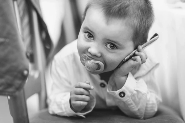 A baby with a dummy holds a mobile phone to his ear and grins cheekily at the camera during a wedding reception. Black and white candid documentary portrait.