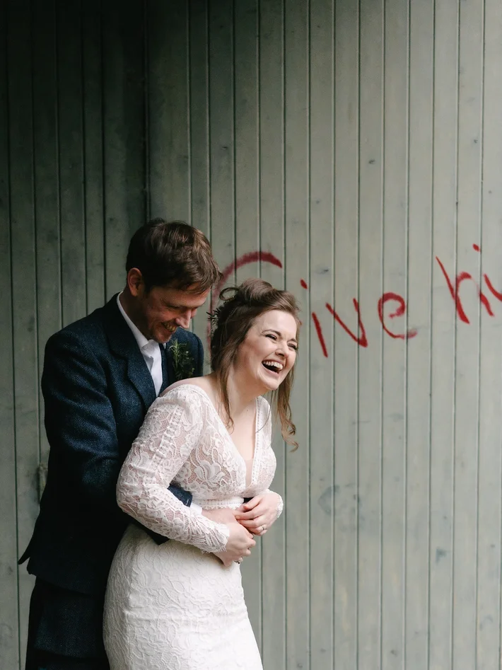 Laughing bride in lace gown embraced by groom in navy suit against weathered wooden wall with red graffiti
