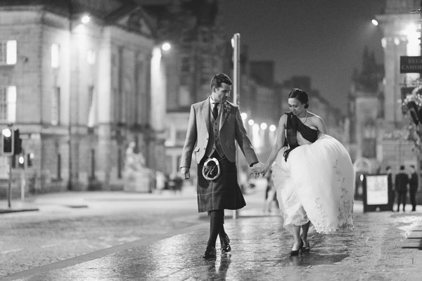 Newlyweds walk hand in hand on a night city street — groom in tartan, bride in white lace