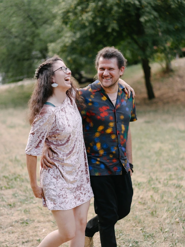 Couple laughing together in field with green trees at outdoor wedding wearing patterned casual clothing.