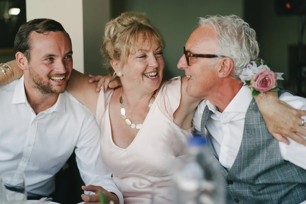 Three guests laughing at the wedding — woman in pearl necklace and pale pink, young man, older man