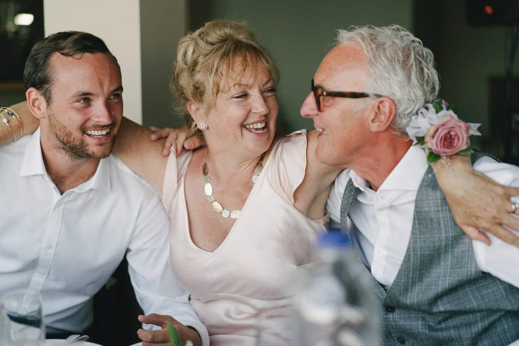 Three guests laughing at the wedding — woman in pearl necklace and pale pink, young man, older man
