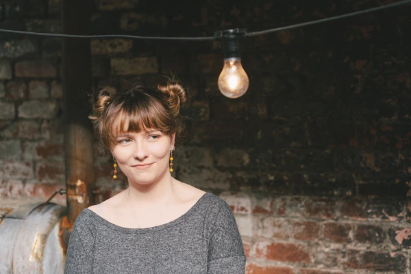 Smiling woman in grey polo, pinned-up hair and yellow earrings, rustic brick background