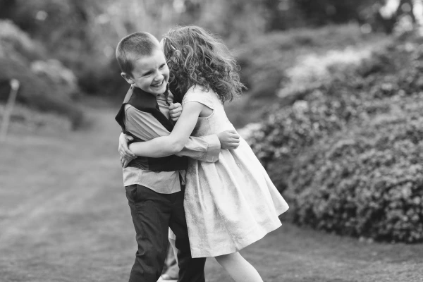 Black and white candid image of two children in formal wedding clothes hugging and laughing together on the garden lawn at the Royal Botanic Garden Edinburgh. Documentary wedding photography.
