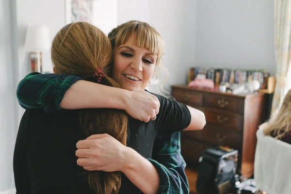 Cheerful woman in green checked shirt hugging another girl with closed eyes, natural light