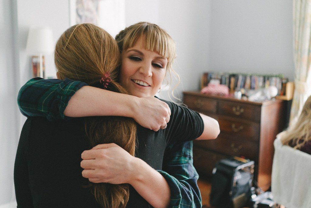Cheerful woman in green checked shirt hugging another girl with closed eyes, natural light