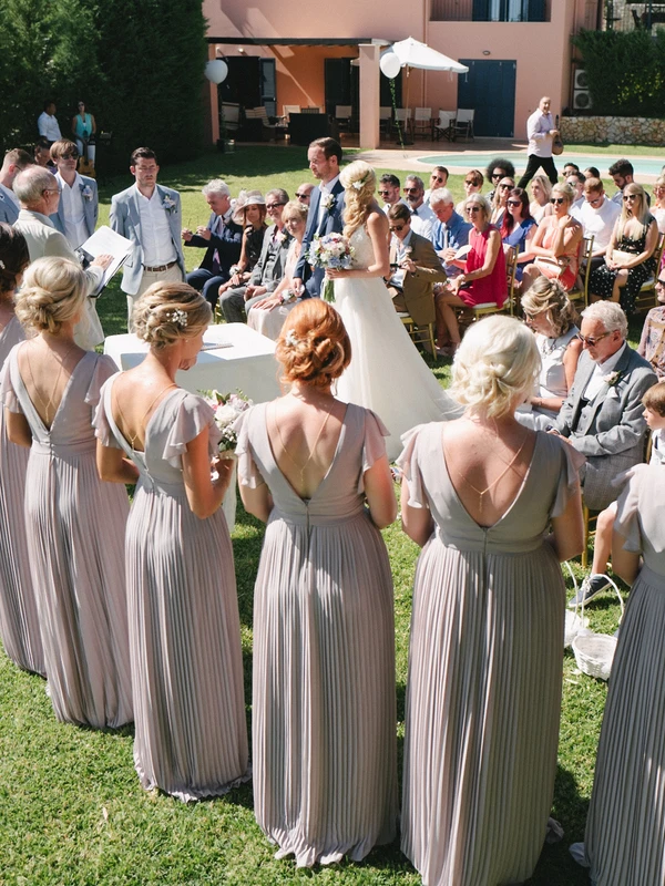 Ceremony from behind the groomsmen in elegant beige — bride seated, guests in sunny garden