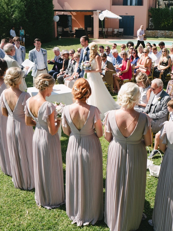 Ceremony from behind the groomsmen in elegant beige — bride seated, guests in sunny garden