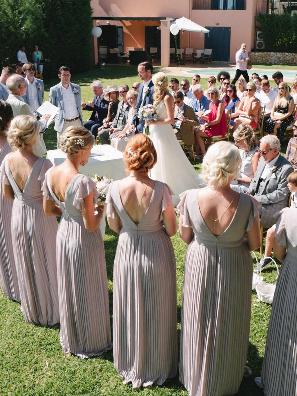 Ceremony from behind the groomsmen in elegant beige — bride seated, guests in sunny garden