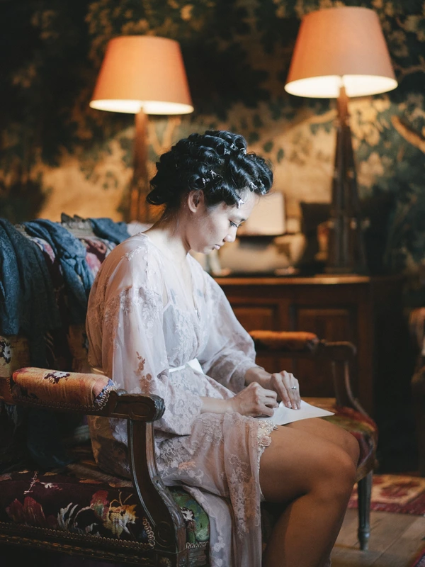 Bride in white lace robe sits in a classic armchair, reading papers — warm lamp light, art deco room