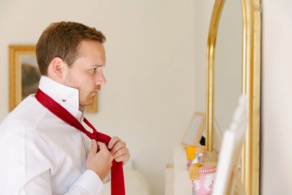 Groom tying a red knit tie in front of a gold-framed mirror during wedding morning preparations. Candid documentary portrait.