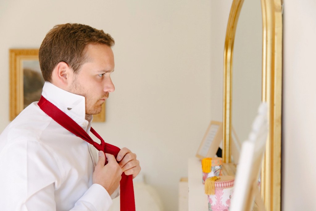 Groom tying a red knit tie in front of a gold-framed mirror during wedding morning preparations. Candid documentary portrait.