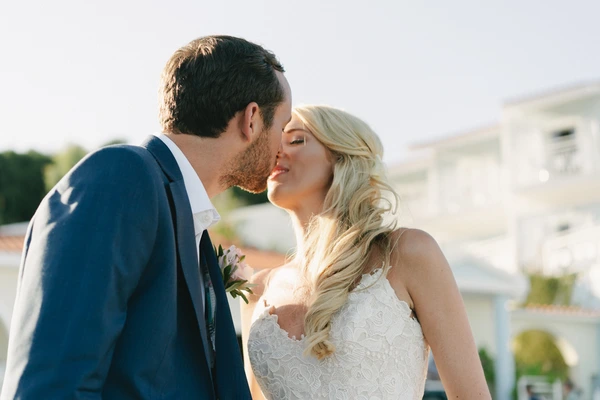 Bride and groom nearly kiss on a sun-drenched terrace — blue suit, white lace, golden afternoon light