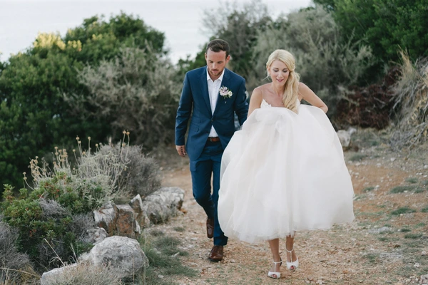 Newlyweds walk a cobbled Mediterranean lane — blue suit, white tulle dress, happy natural moment