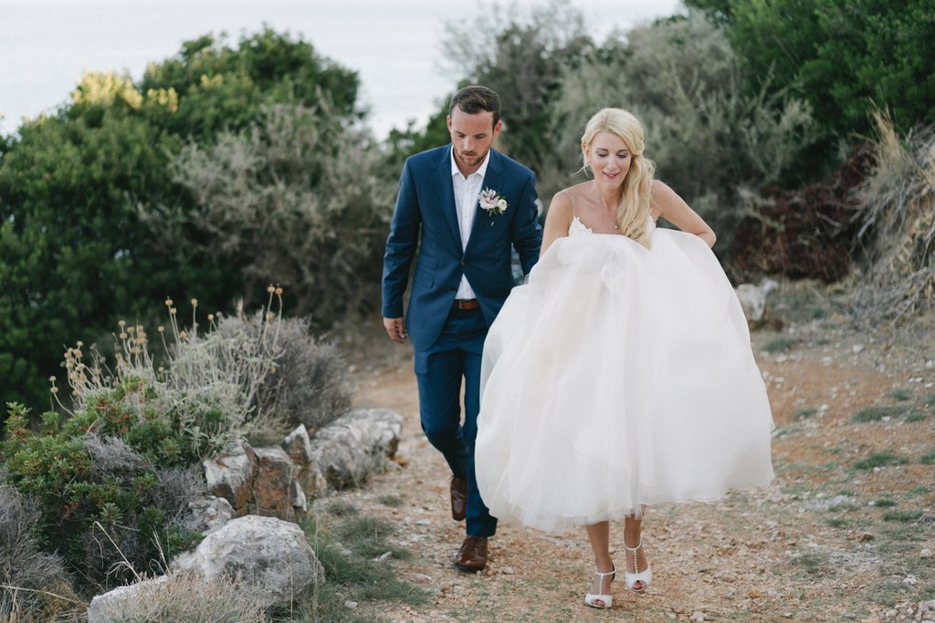 Newlyweds walk a cobbled Mediterranean lane — blue suit, white tulle dress, happy natural moment
