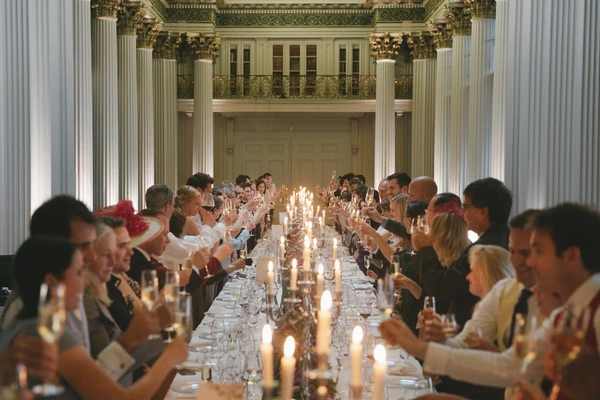 Wedding guests at a ceremonial banquet — golden columns, decorated ceiling, raised glasses