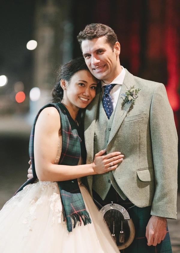 Couple embrace in evening light — groom in beige suit and green scarf, bride in lace and tulle