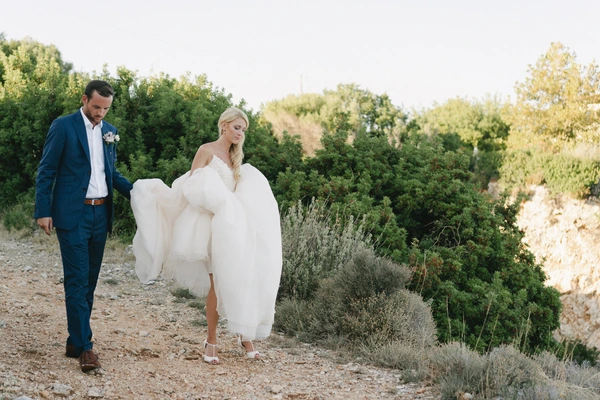 Groom in blue suit leads the bride whose white dress billows in the wind along rocky terrain