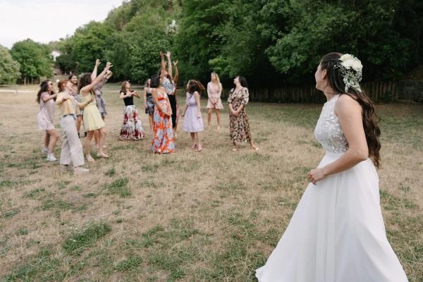Bride preparing to toss bouquet to female guests standing in field during outdoor wedding celebration.