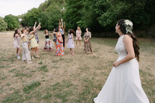 Bride preparing to toss bouquet to female guests standing in field during outdoor wedding celebration.