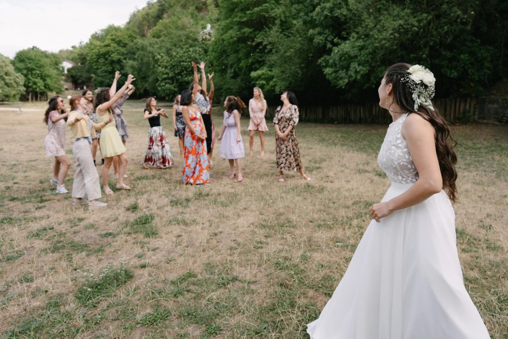 Bride preparing to toss bouquet to female guests standing in field during outdoor wedding celebration.