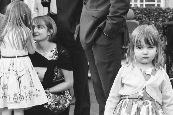 Black and white candid image of a young girl with a serious expression among wedding guests, while a woman crouches to speak to a child beside her. Documentary wedding photography.