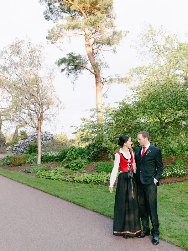 The couple stand close together in the garden, the bride in her Norwegian bunad looking up at the groom as they talk softly. Candid documentary wedding portrait at the Royal Botanic Garden Edinburgh.