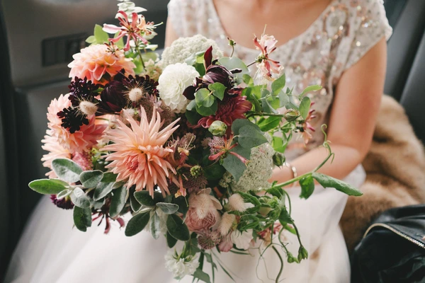 Bride in white lace holds a wildflower bouquet with orange dahlias, burgundy roses and whites