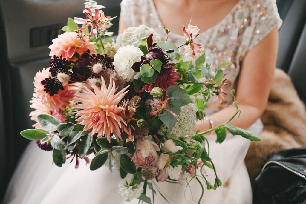 Bride in white lace holds a wildflower bouquet with orange dahlias, burgundy roses and whites