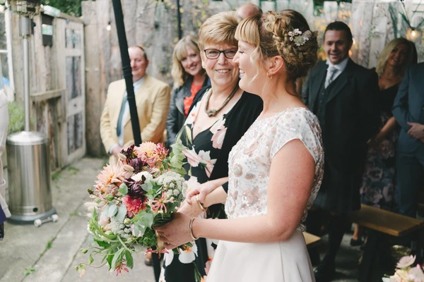 Bride in white lace with bouquet talking to a smiling woman in black — guests in garden