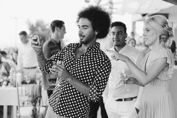 Man in spotted shirt speaks with a microphone, smiling blonde woman beside him, guests in white room