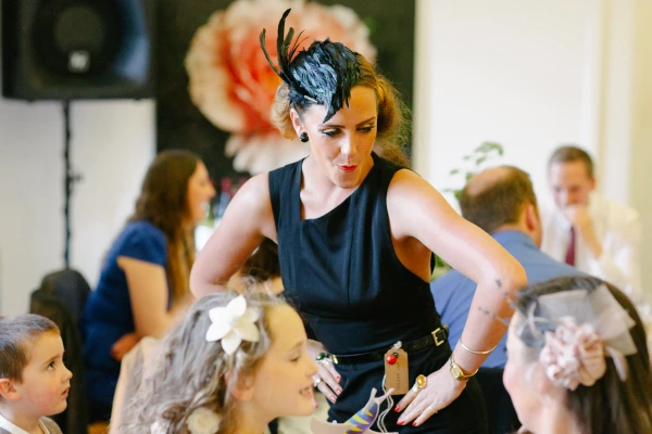 A woman in a black dress and feathered fascinator leans over to talk to children at a wedding reception, a large paper flower decoration visible behind her. Candid documentary photograph.