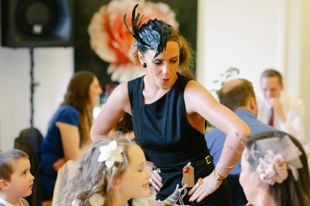 A woman in a black dress and feathered fascinator leans over to talk to children at a wedding reception, a large paper flower decoration visible behind her. Candid documentary photograph.