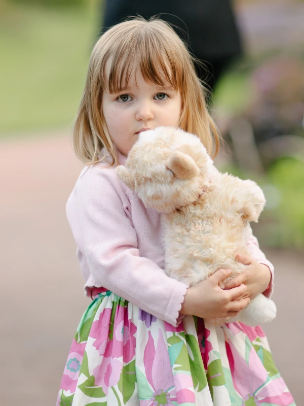 A young girl in a pink cardigan and floral dress hugs a fluffy stuffed animal and gazes directly into the camera at the Royal Botanic Garden Edinburgh. Candid documentary wedding portrait.