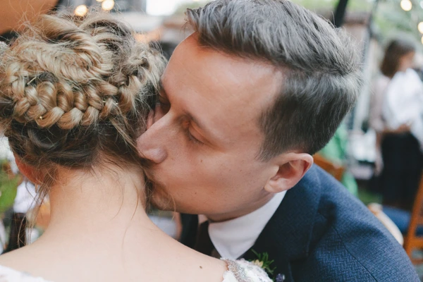 Groom kisses the bride on the neck — braided updo, decorative lanterns, guests at the wedding