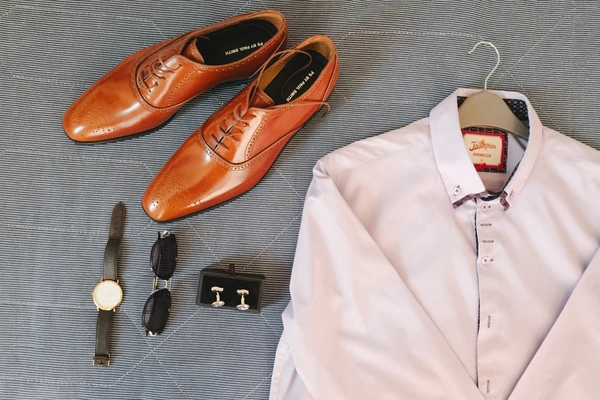 Groom's flat lay — pink shirt, brown Oxford shoes, black watch, silver cufflinks and sunglasses