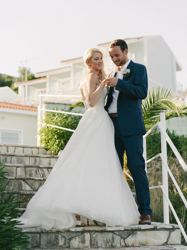 Groom in dark blue suit presents bouquet to joyful bride on a terrace — white building and greenery