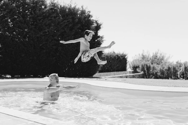 Boy jumps into the pool while a woman smiles and watches from the water — green trees behind