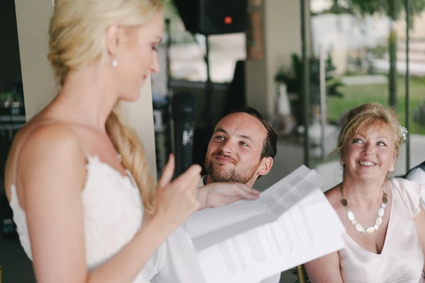 Bride in white smiles toward a man and woman who watch her with emotion at the outdoor venue