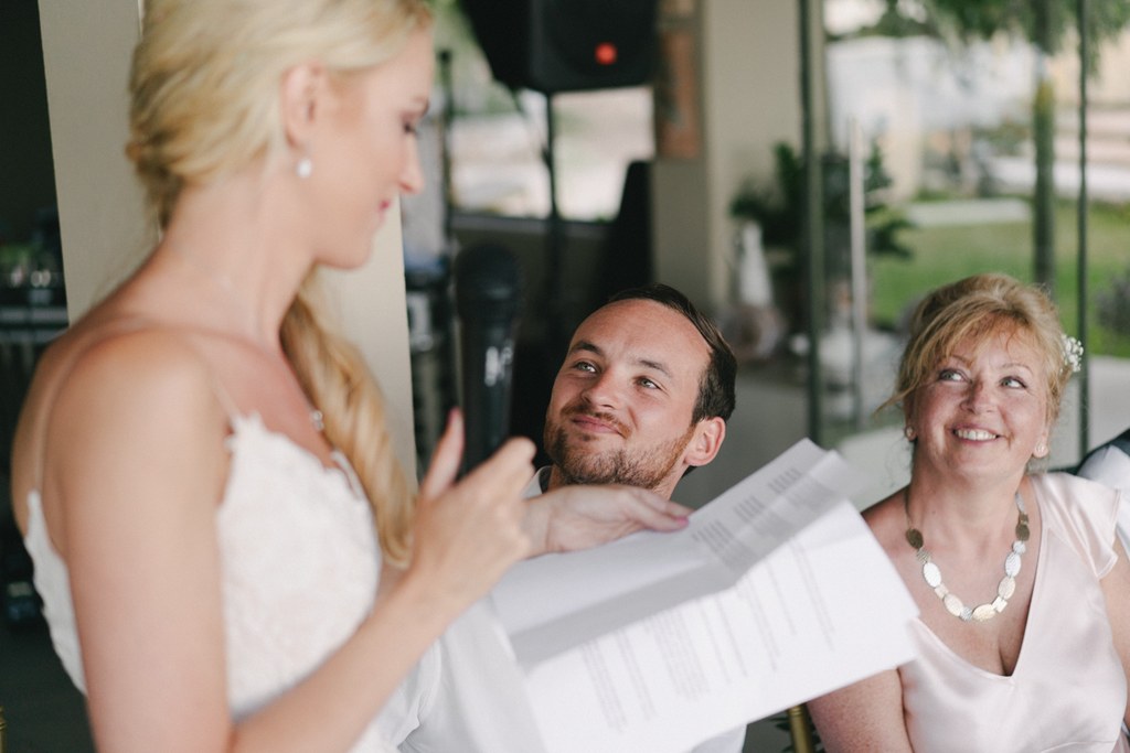 Bride in white smiles toward a man and woman who watch her with emotion at the outdoor venue
